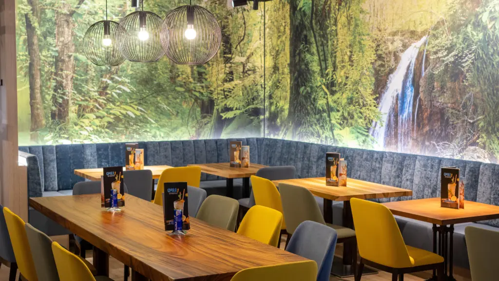 Gemütlicher Sitzbereich mit Holztischen und großem Dschungel-Wandbild mit Wasserfall im Restaurant CHILLY in St. Johann im Pongau