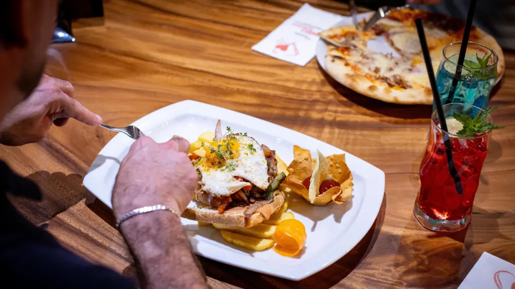 Gast isst ein Gericht mit Spiegelei und Pommes, dazu bunte Cocktails und Pizza auf dem Tisch im Restaurant CHILLY in St. Johann im Pongau